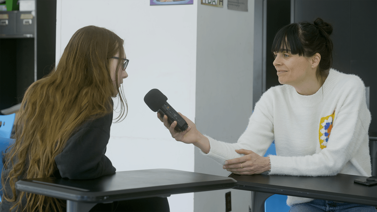 Young interviewer holding a microphone across a desk from a woman in a white sweater in a classroom setting